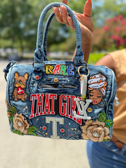A hand holds a heavily embellished blue denim handbag outdoors. The bag features embroidered patches, rhinestone lettering reading 'THAT GIRL' and 'RARE', floral appliqués, a dog patch, and a woman's face patch.